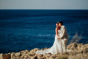 A couple in wedding attire embraces on rocky coastal cliffs overlooking a deep blue ocean at sunset.