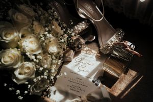 Wedding details flatlay with white roses, invitation, perfume bottle and heels in moody lighting.