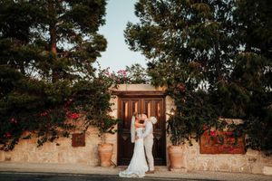 A couple stands in a romantic garden setting with stone walls and flowering trees creating an elegant backdrop.