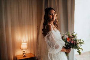 Bride in white robe holding bouquet near window with warm lamp lighting.