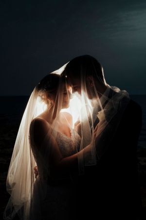 Dramatic backlit silhouette of couple embracing under wedding veil with glowing light effect.