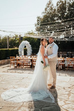 A romantic outdoor wedding ceremony setup with string lights, rustic wooden chairs, and a white floral arch on a stone patio.