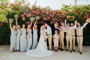 Wedding party in light gray dresses and tan suits poses together in front of vibrant red flowering bushes.