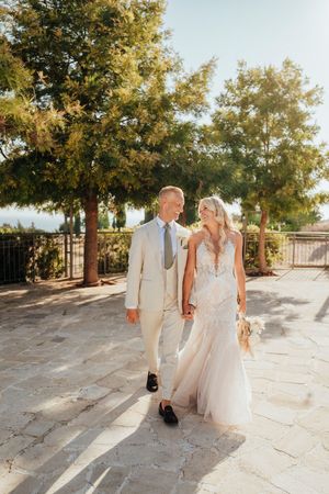 Newlywed couple walks hand in hand along a sunlit stone path with trees casting dappled shadows on a bright summer day.