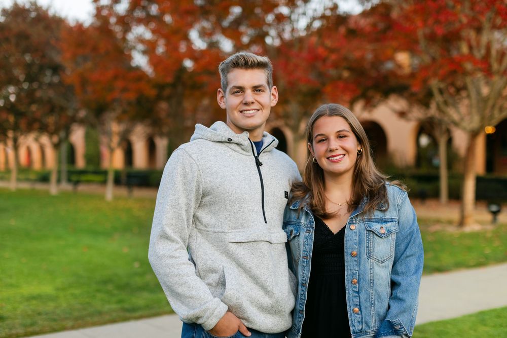 Two smiling young adults posing outdoors on a campus with vibrant autumn foliage in the background.