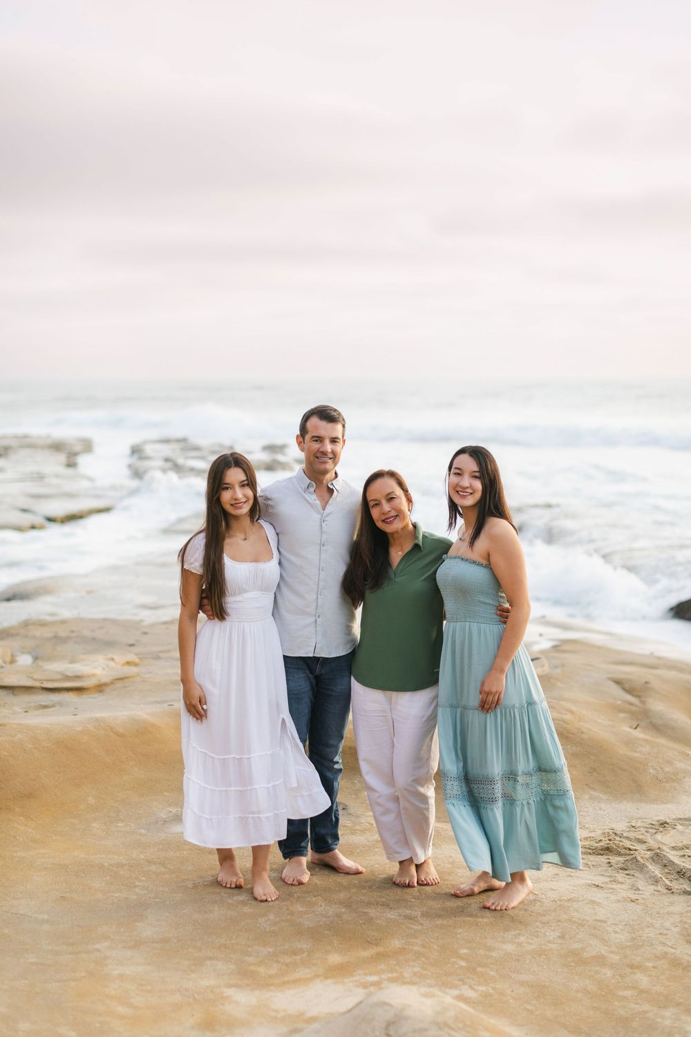 Family of four smiling together on rocky beach shoreline, wearing casual summer outfits with ocean waves behind them.