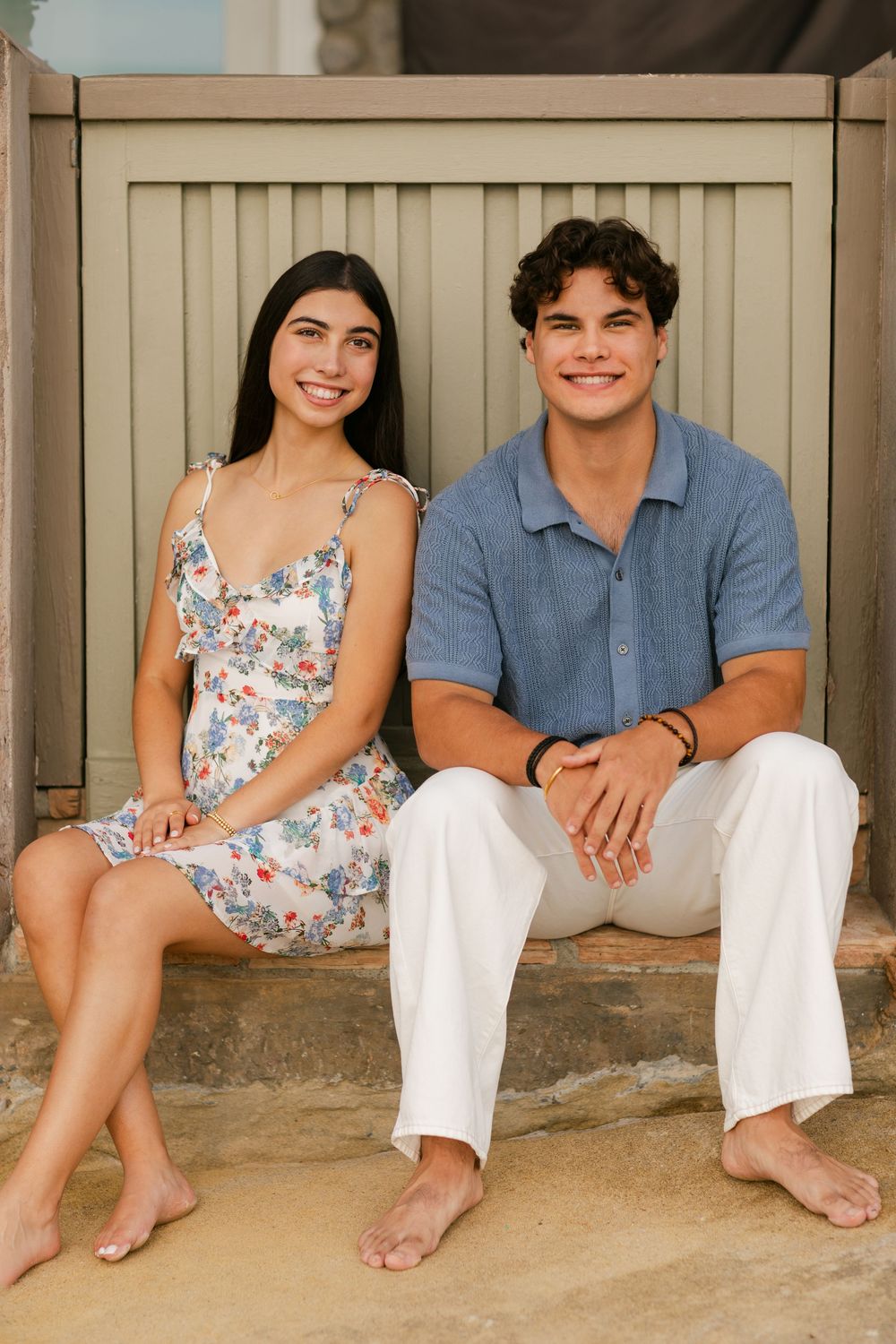 A young couple smiling and sitting barefoot on steps, wearing casual summer outfits outdoors.