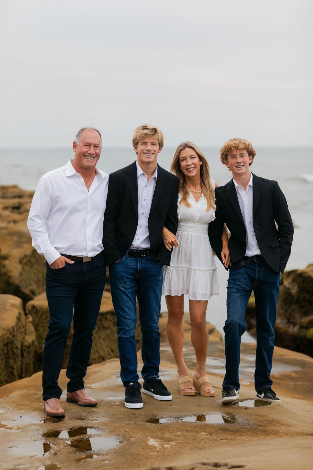 Family of four posing on rocky beach with ocean backdrop, wearing smart casual attire including blazers and white outfits.