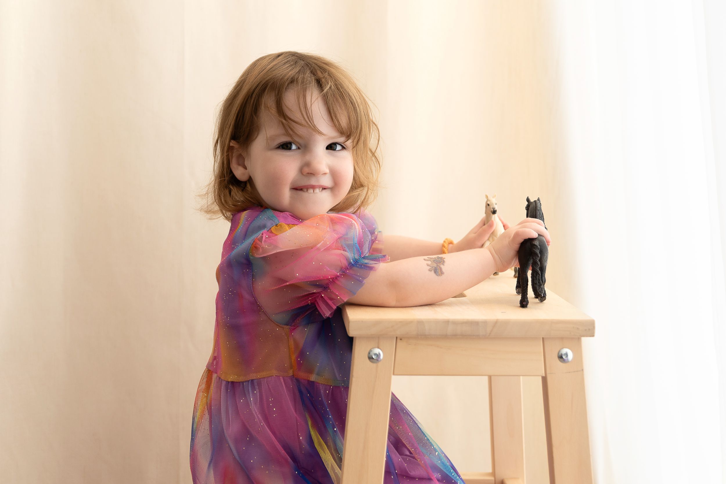 A small child in a colorful dress plays with a toy figure while standing at a wooden table against a beige wall.