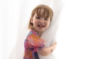 Joyful child in colorful tie-dye shirt peeking and laughing from behind white curtain.