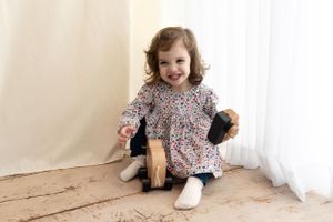 Small child wearing floral print dress sits on wooden floor holding plush toys and laughing joyfully.