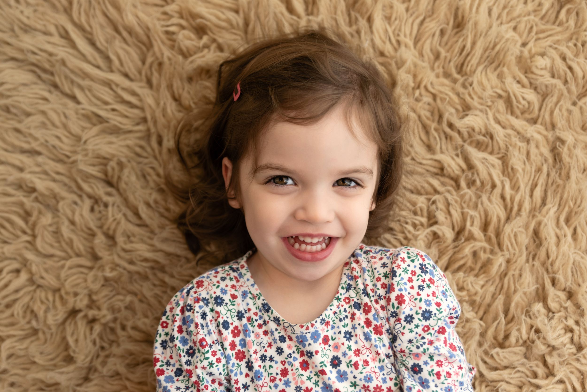 Child lying on fluffy beige carpet wearing a floral print dress with a bright smile.