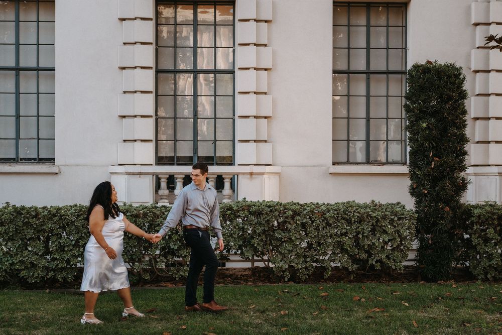 A couple walks hand in hand along a white building with tall windows and manicured hedges.