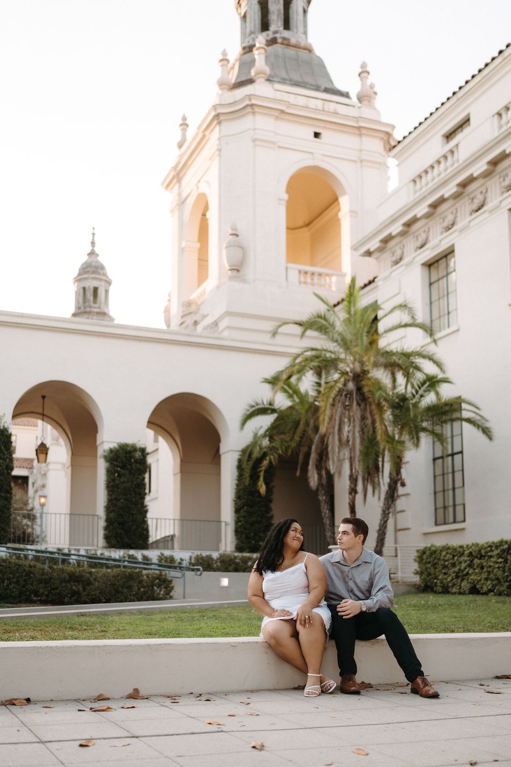 A couple sits together on white stone steps at an elegant Spanish colonial style building with arches and palm trees.