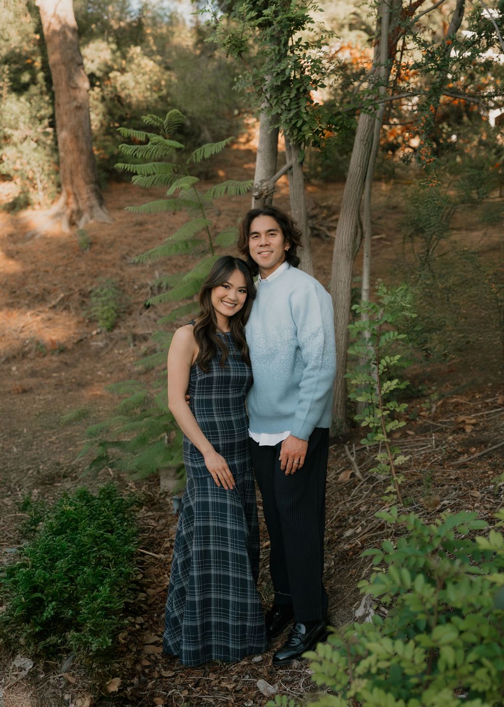 A couple poses together in a wooded outdoor setting during golden hour, wearing a plaid dress and blue shirt.