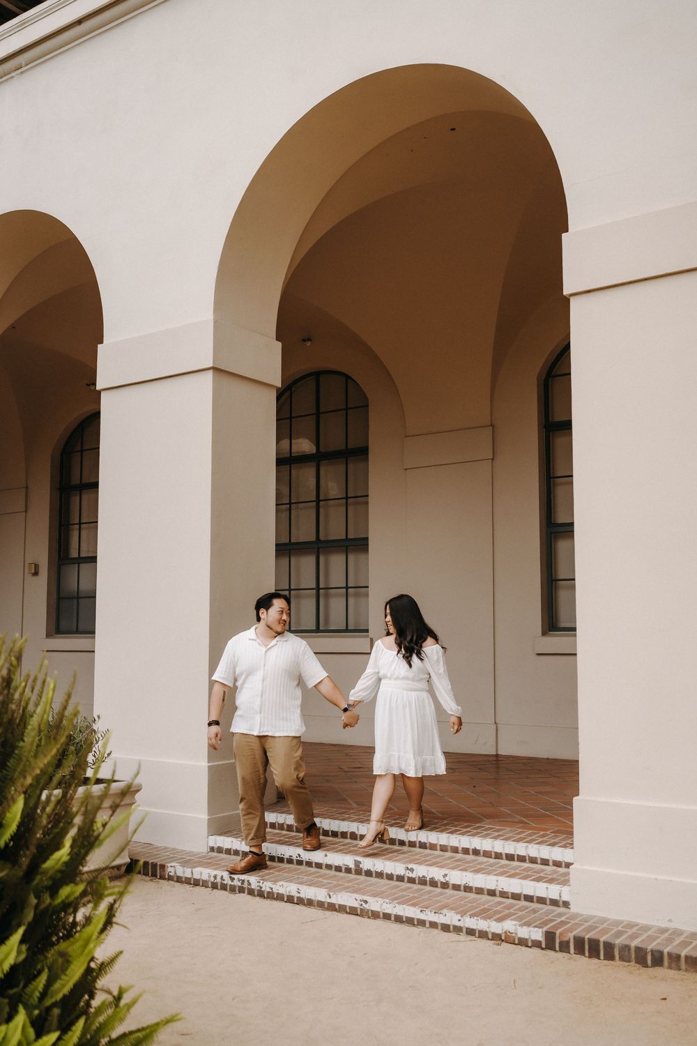A couple walks together along a white arched colonnade with Spanish-style architecture in warm sunlight.
