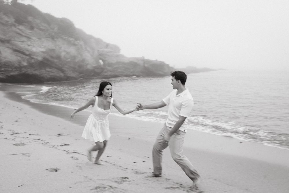 Black and white photo of couple dancing playfully on a foggy beach.