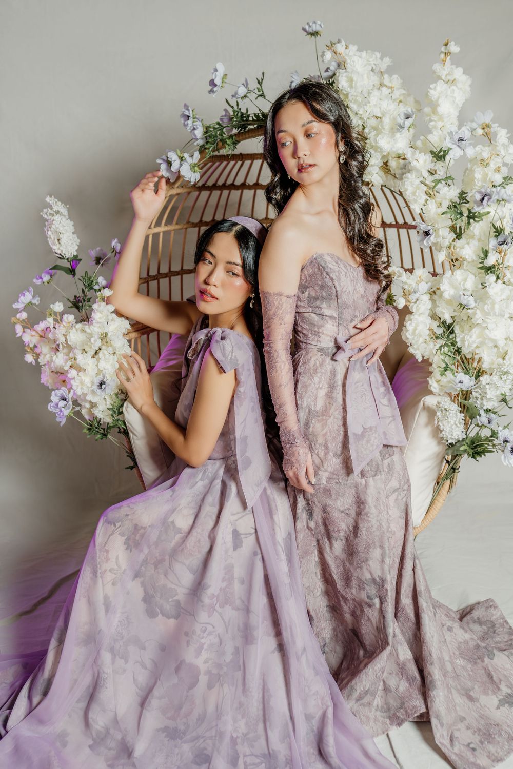 Elegant fashion photo with two models in flowing lavender gowns posing with white floral arrangements and birdcage decor.