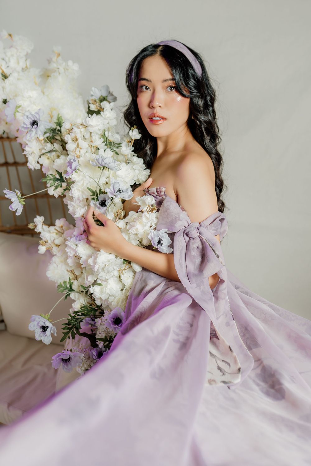 Romantic portrait in a flowing lilac off-shoulder gown with white floral arrangements and soft lighting.