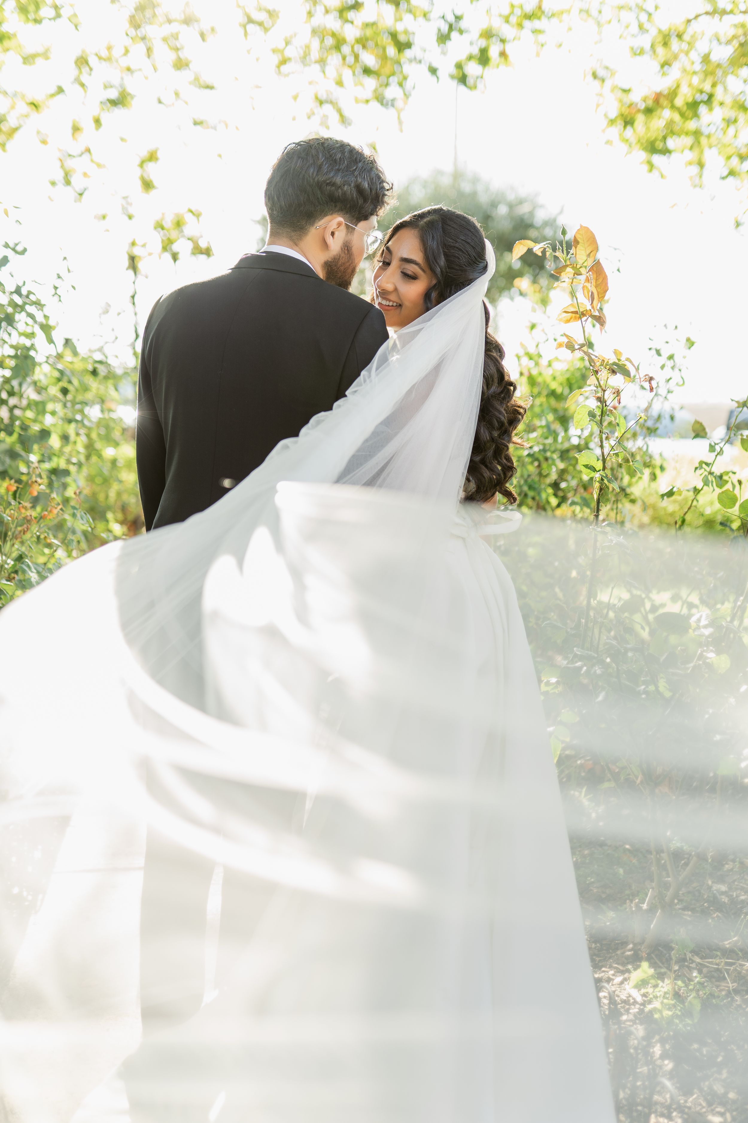 A romantic wedding couple embraces outdoors as the bride's flowing white veil catches the sunlight in a dreamy scene.