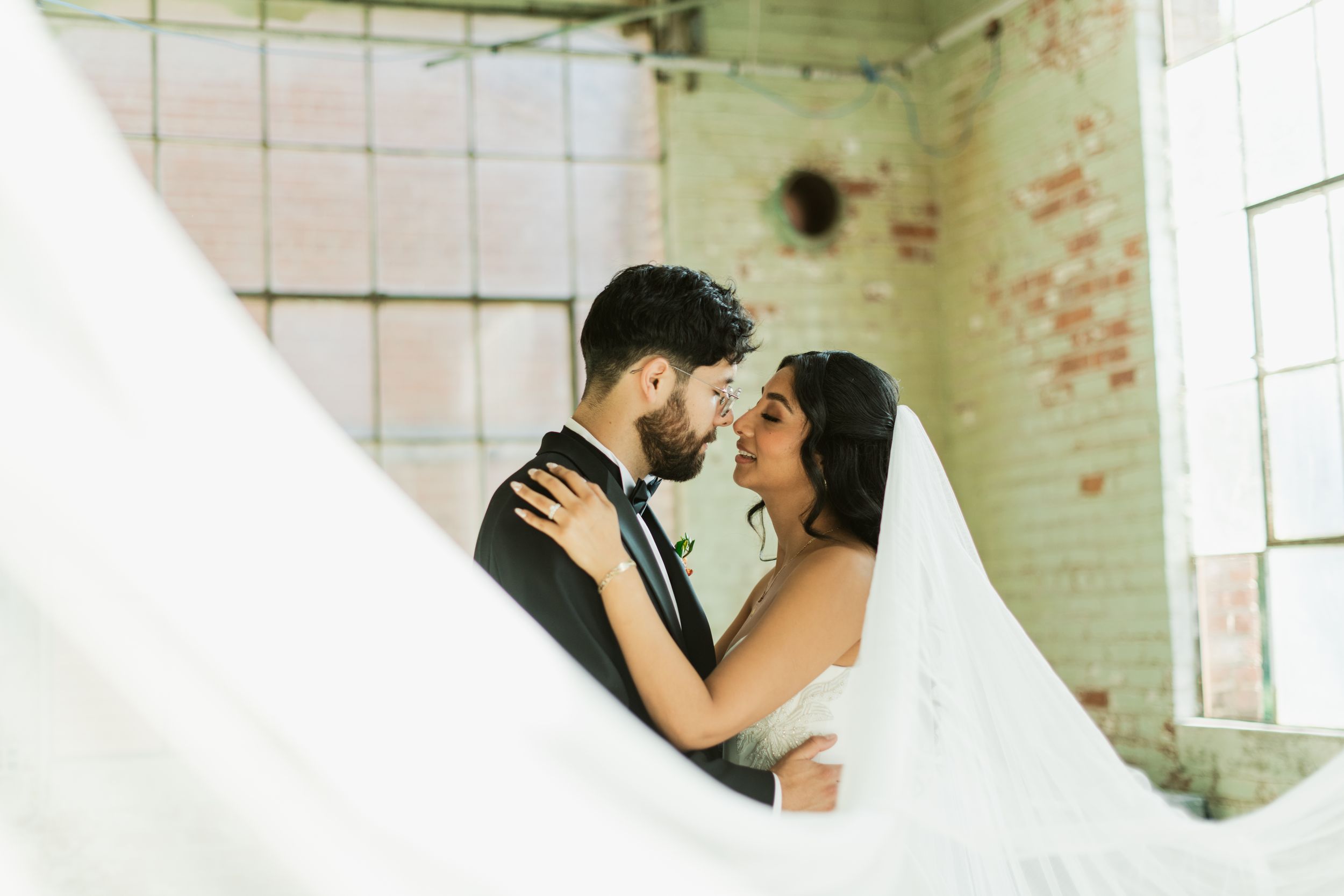 A romantic wedding couple embraces and kisses in a rustic brick room with natural window lighting.