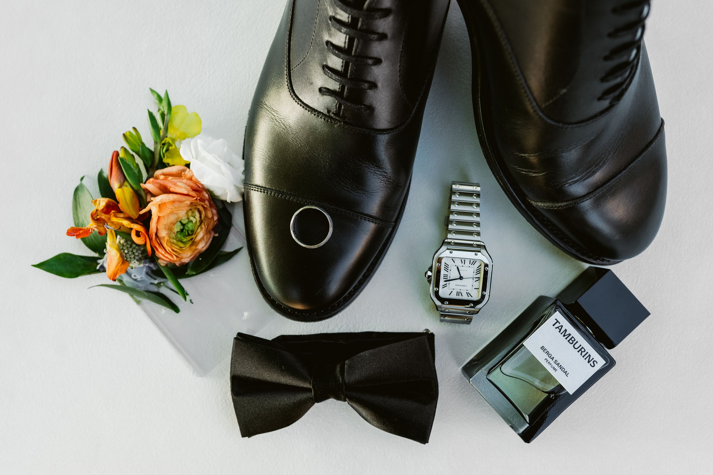 Black dress shoes arranged with bow tie and floral boutonniere on white background for formal occasion styling.