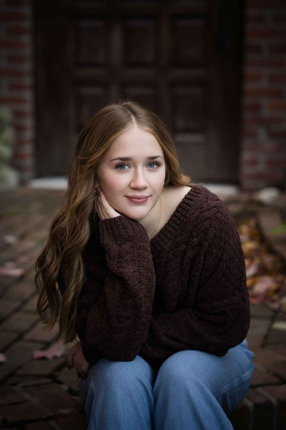 Young woman in brown knit sweater and jeans sits on brick steps, smiling warmly with hands near face.