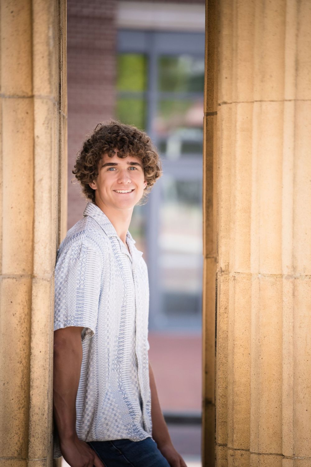 Smiling young man with curly hair leaning against stone columns in a white patterned shirt outdoors.