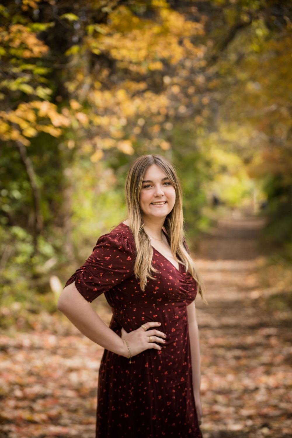 Young woman in burgundy floral dress poses on autumn forest path with colorful fall foliage backdrop.