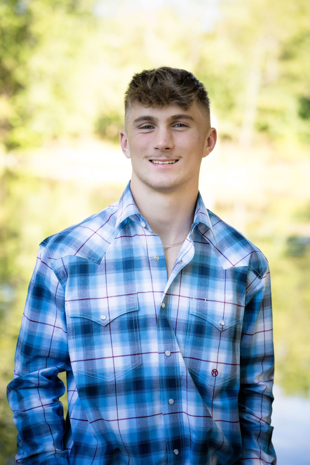 Young man smiling outdoors in blue plaid shirt with nature background.