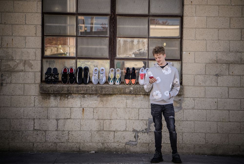 Young man in hoodie and ripped jeans holding phone, standing by brick wall with sneaker collection on windowsill.