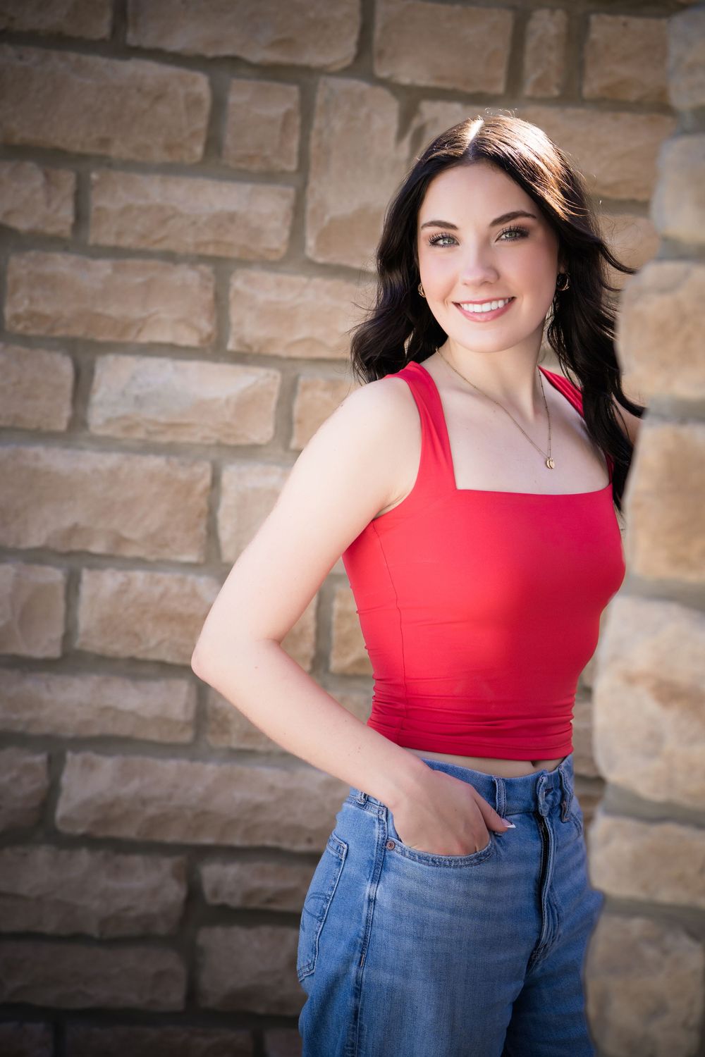Smiling young woman in red crop top and jeans posing confidently against a rustic stone wall.