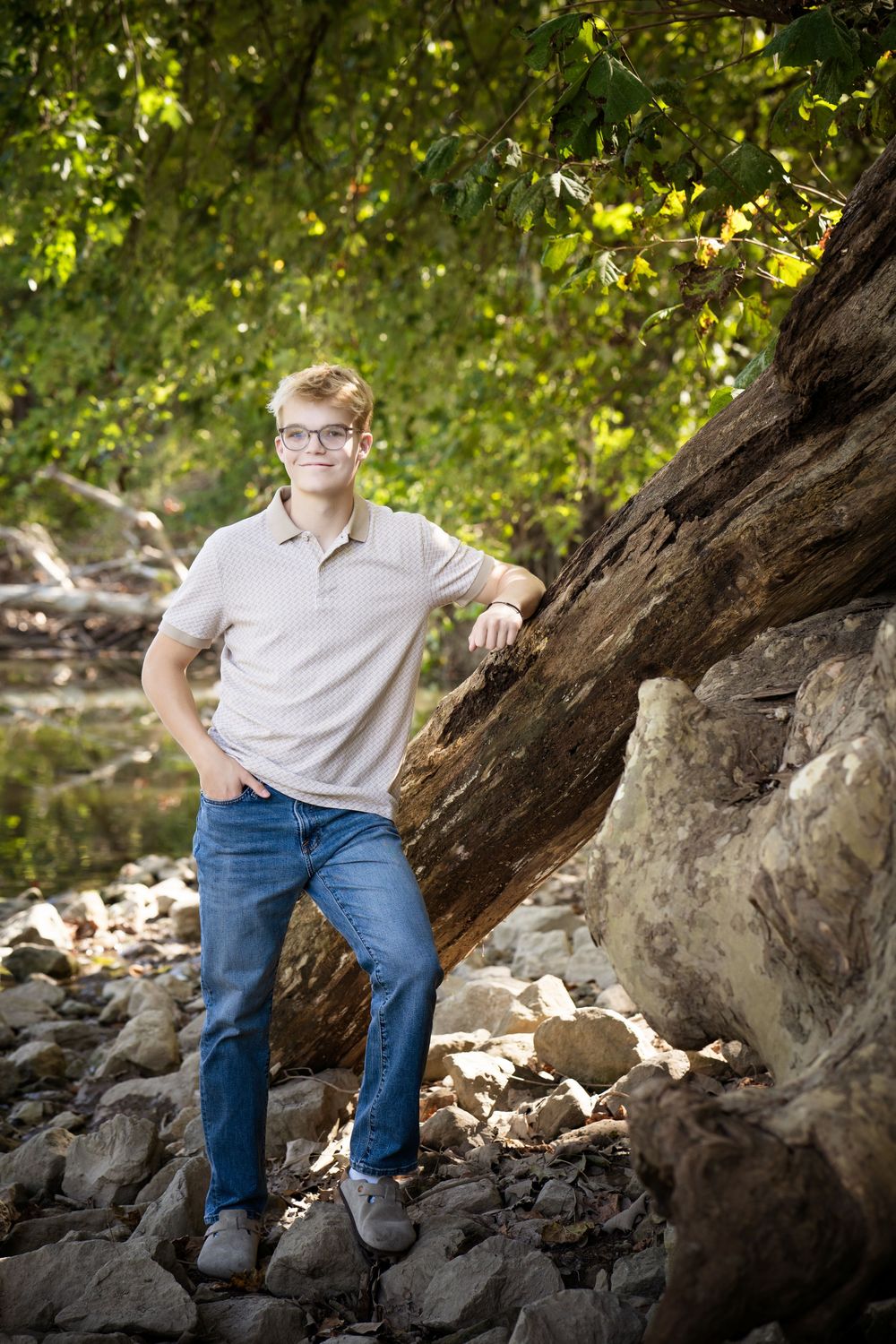 Teen boy in glasses and jeans leans on a fallen log among rocks in a lush green outdoor setting.