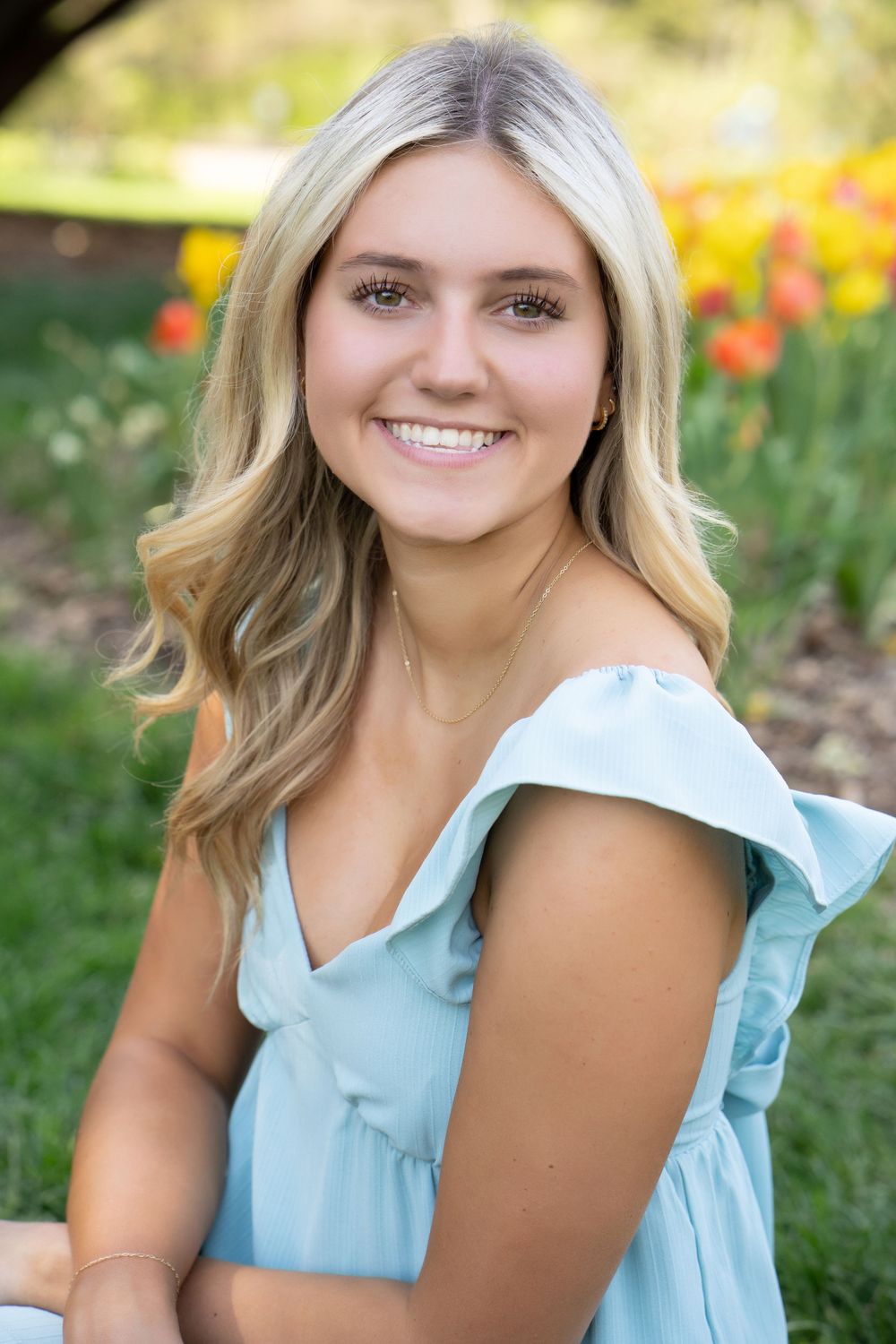 Young blonde woman in light blue off-shoulder dress smiling outdoors with colorful tulips in background.