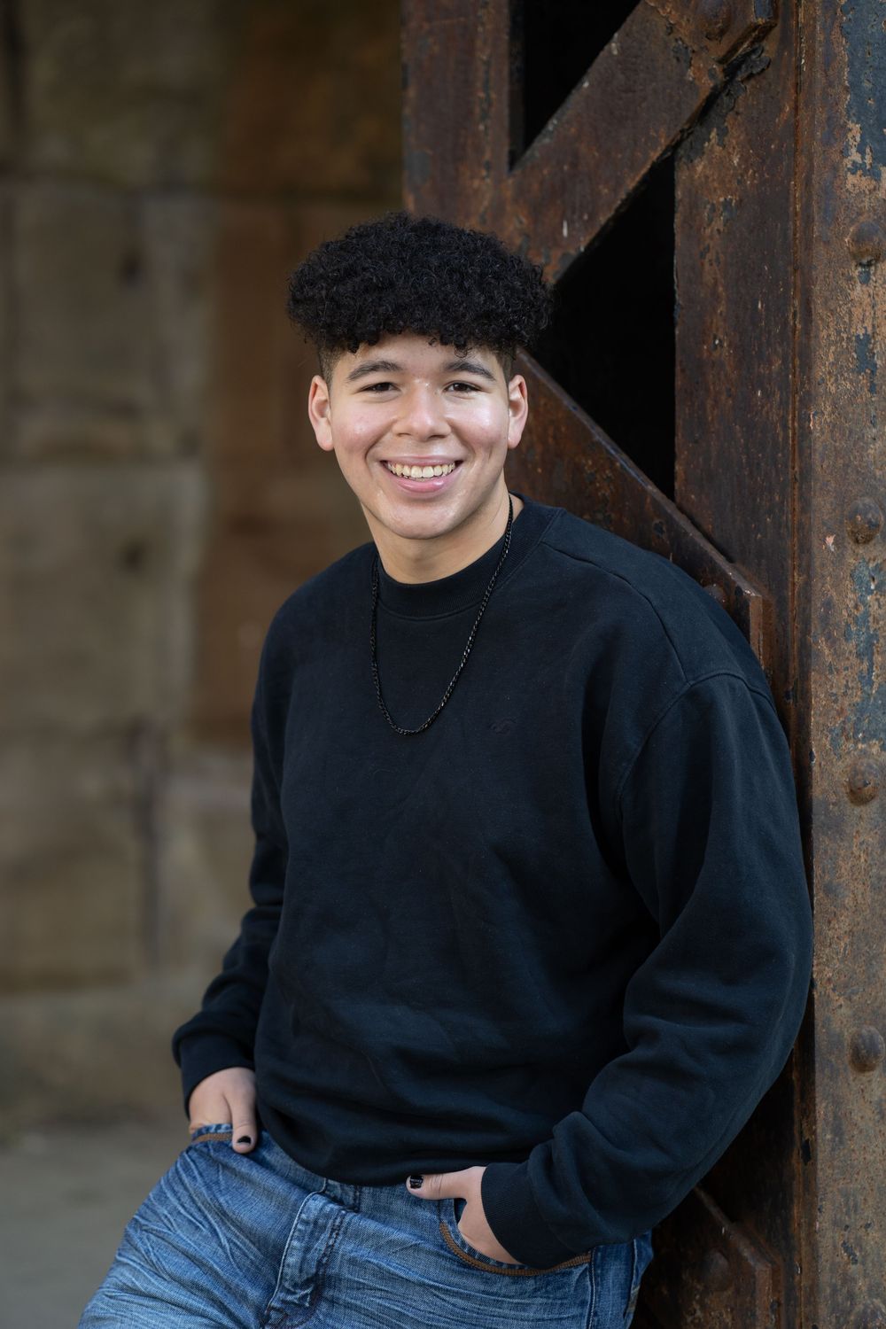 Smiling young man in black sweatshirt and jeans leaning against rustic metal door outdoors.