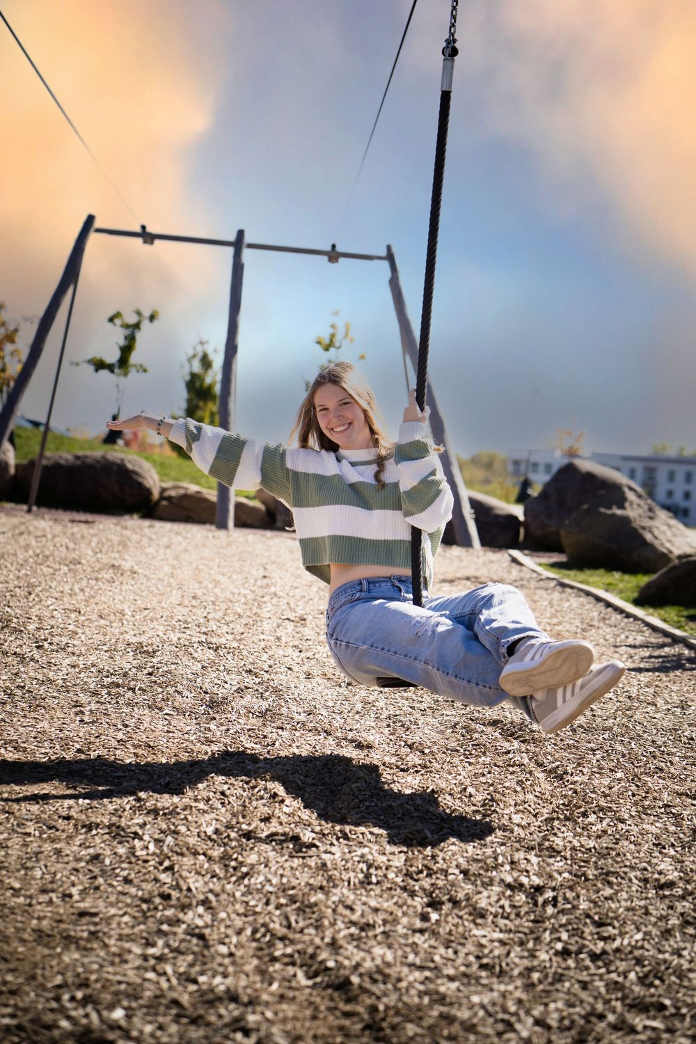 Smiling young woman in striped sweater and jeans sitting on a playground swing on a sunny day.