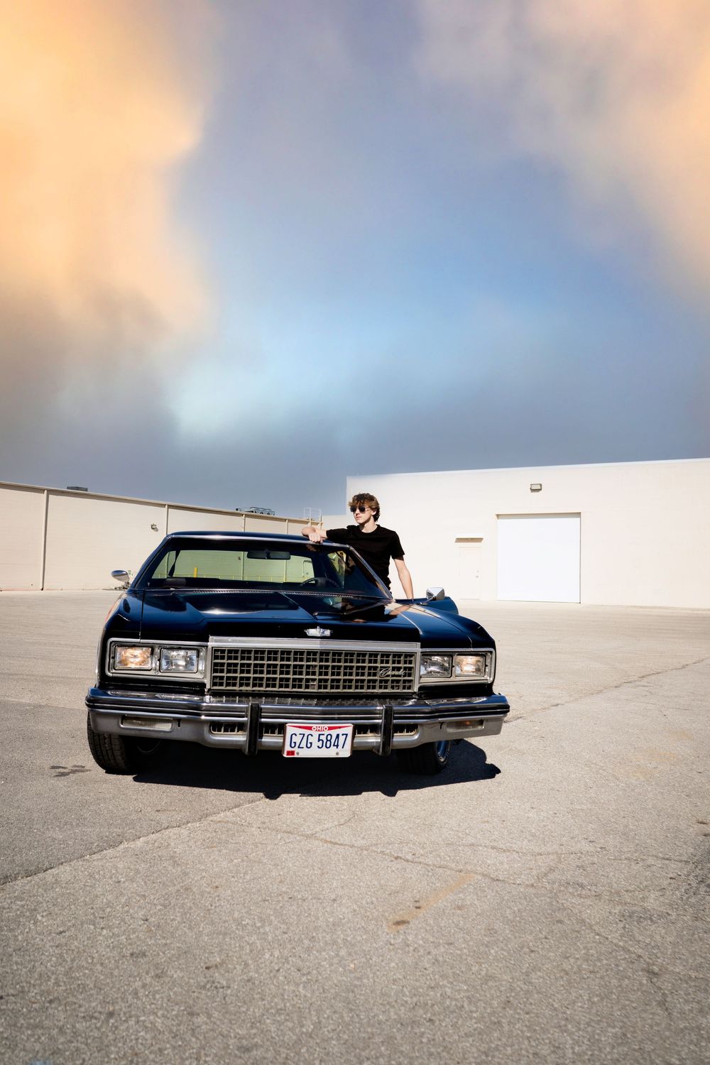 A young man leans on a classic black Chevrolet in an empty parking lot under a dramatic cloudy sunset sky.