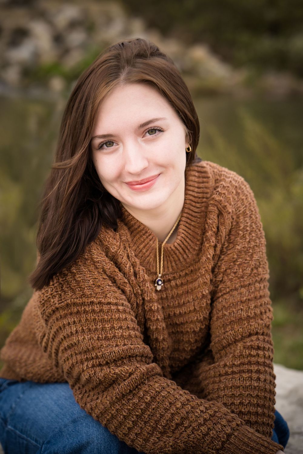 Young woman with brown hair smiling warmly, wearing a brown knit sweater and jeans, seated outdoors.