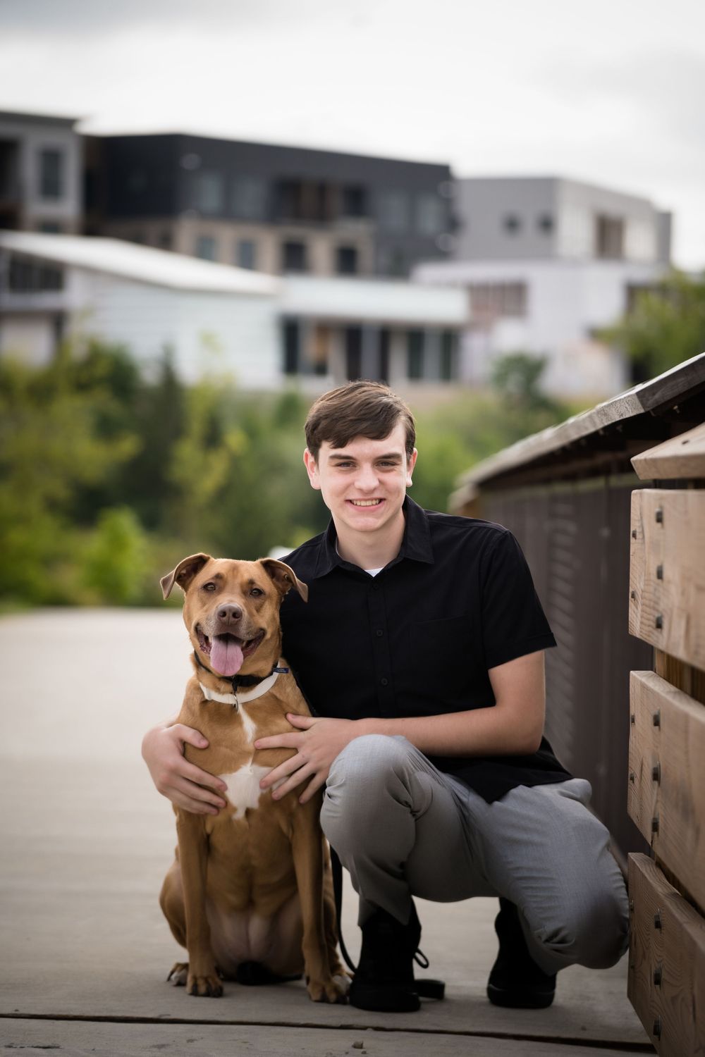 Young man in black shirt smiling with brown pit bull dog on wooden boardwalk near modern buildings.
