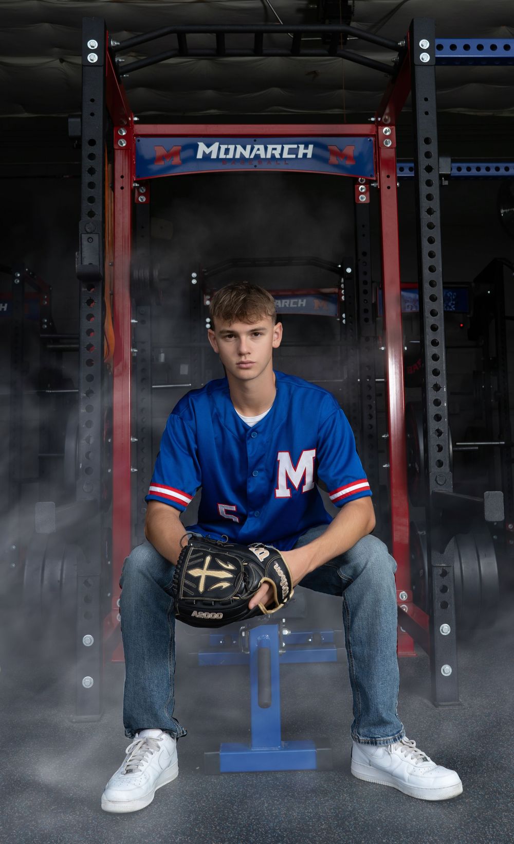 Teen baseball player in blue Monarch jersey holds glove while sitting in front of a power rack in a gym.