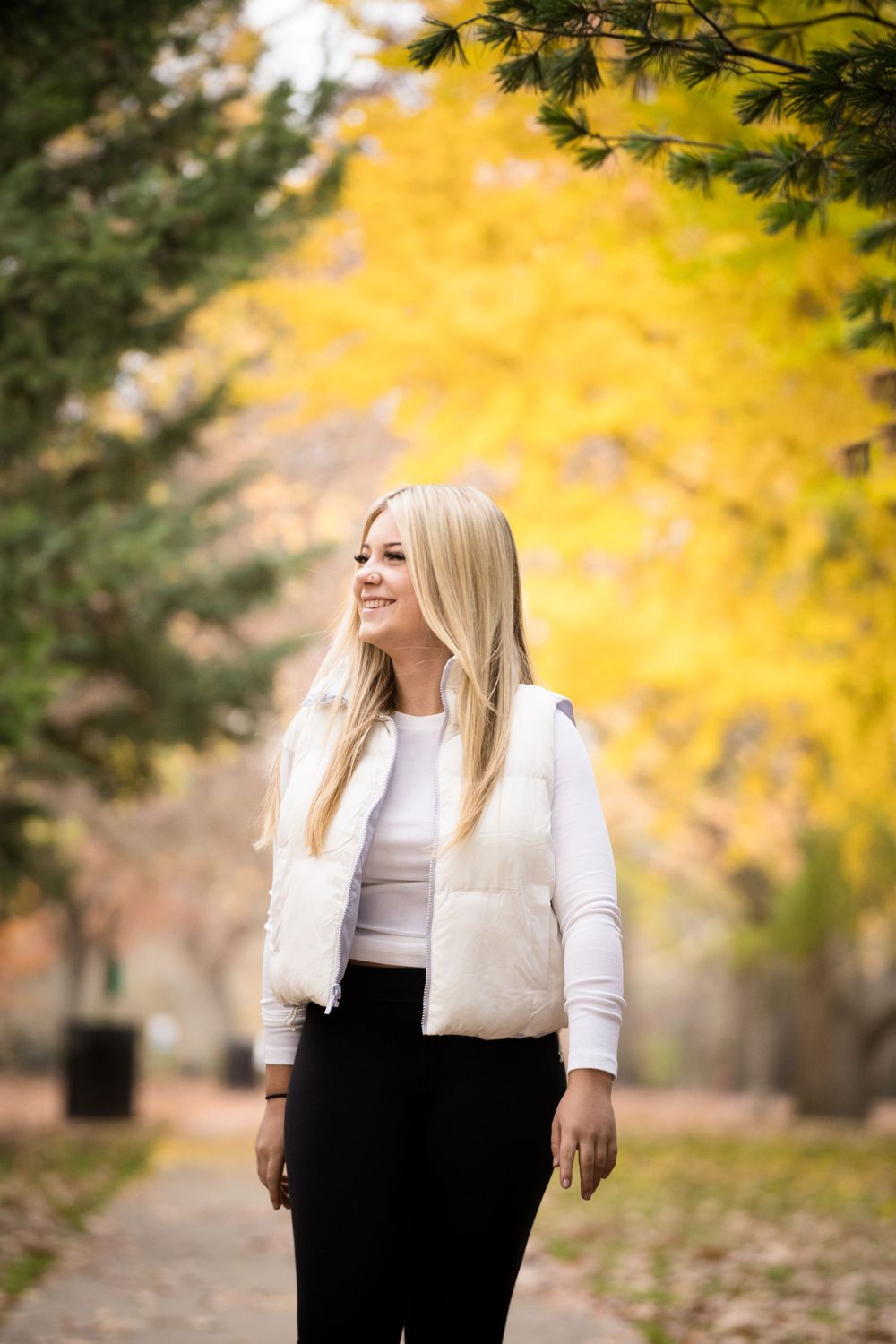 Blonde woman in white puffer vest and black pants smiling outdoors among vibrant yellow autumn trees.