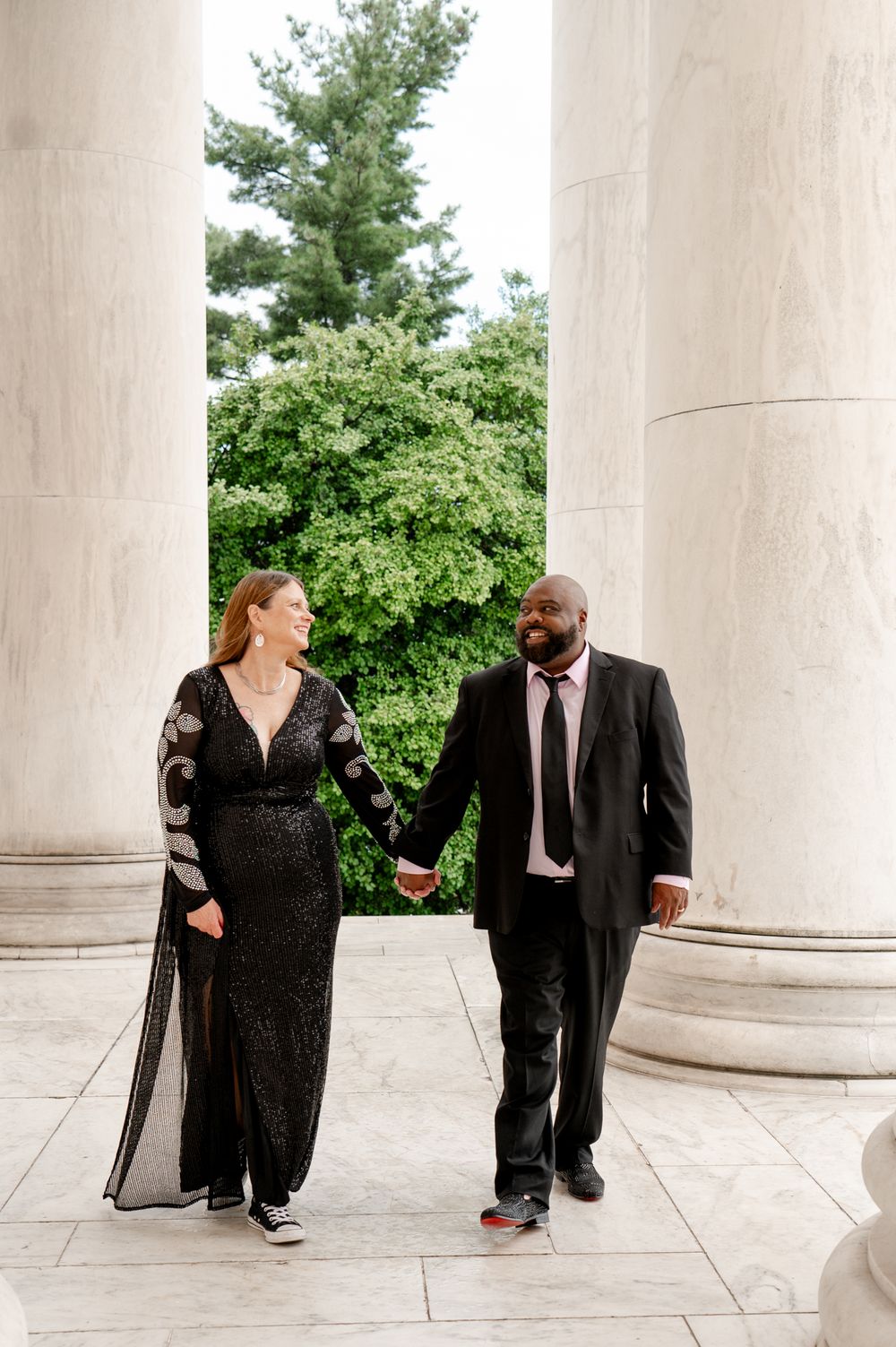 A couple in formal attire holding hands while walking between elegant white marble columns with greenery in background.