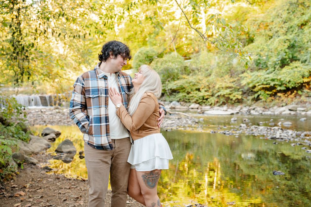 A romantic couple embraces beside a tranquil creek surrounded by autumn foliage on a golden afternoon.