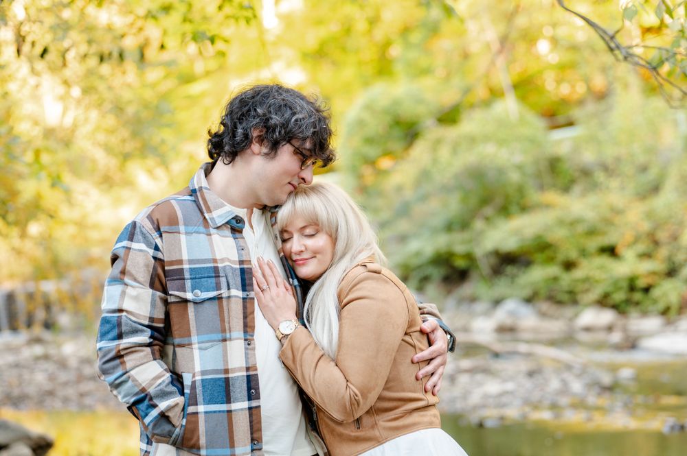 A romantic couple embraces beside a tranquil creek surrounded by autumn foliage on a golden afternoon.
