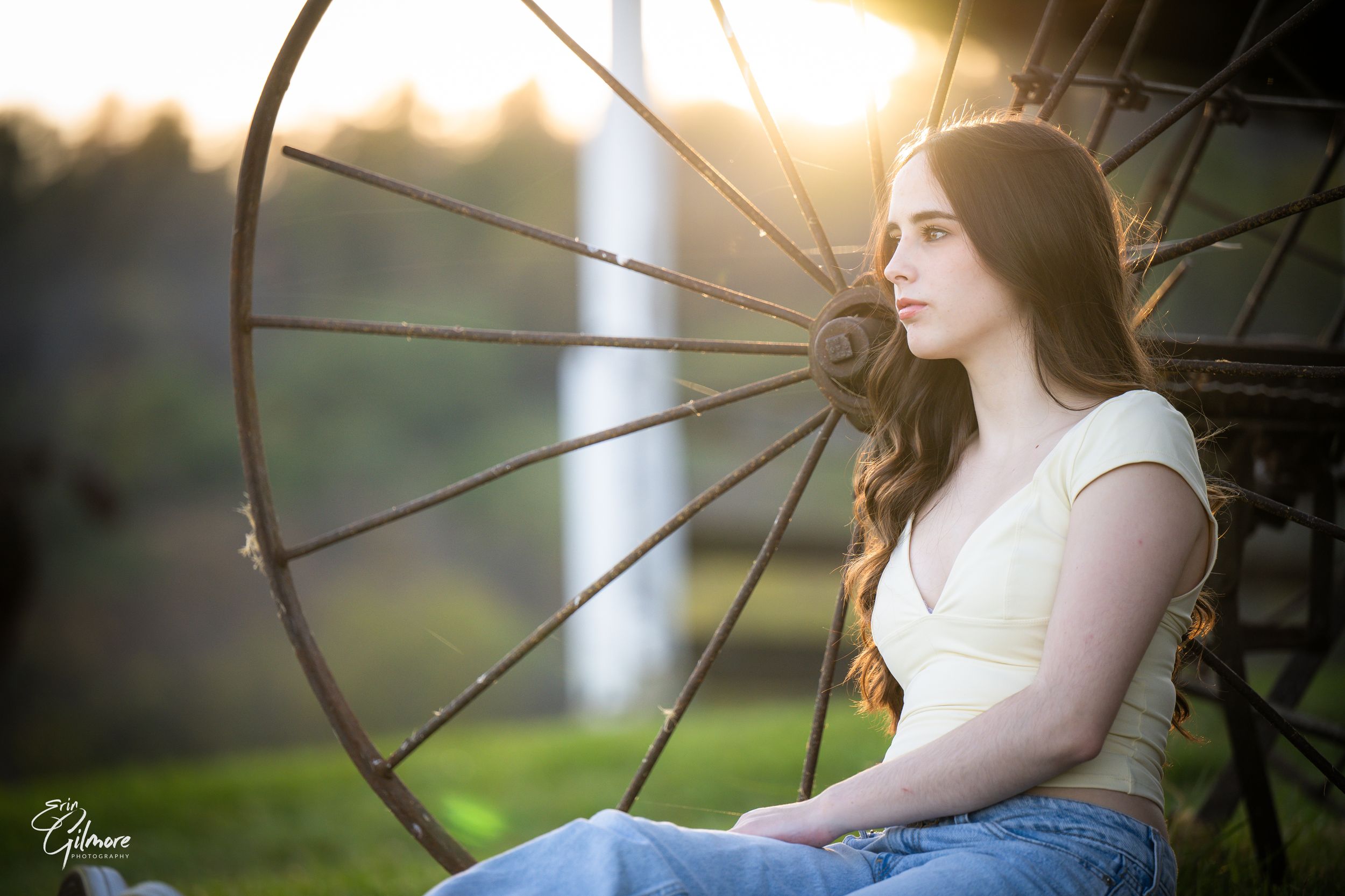 Young woman in yellow top and jeans sits pensively beside a rustic wagon wheel at golden hour.
