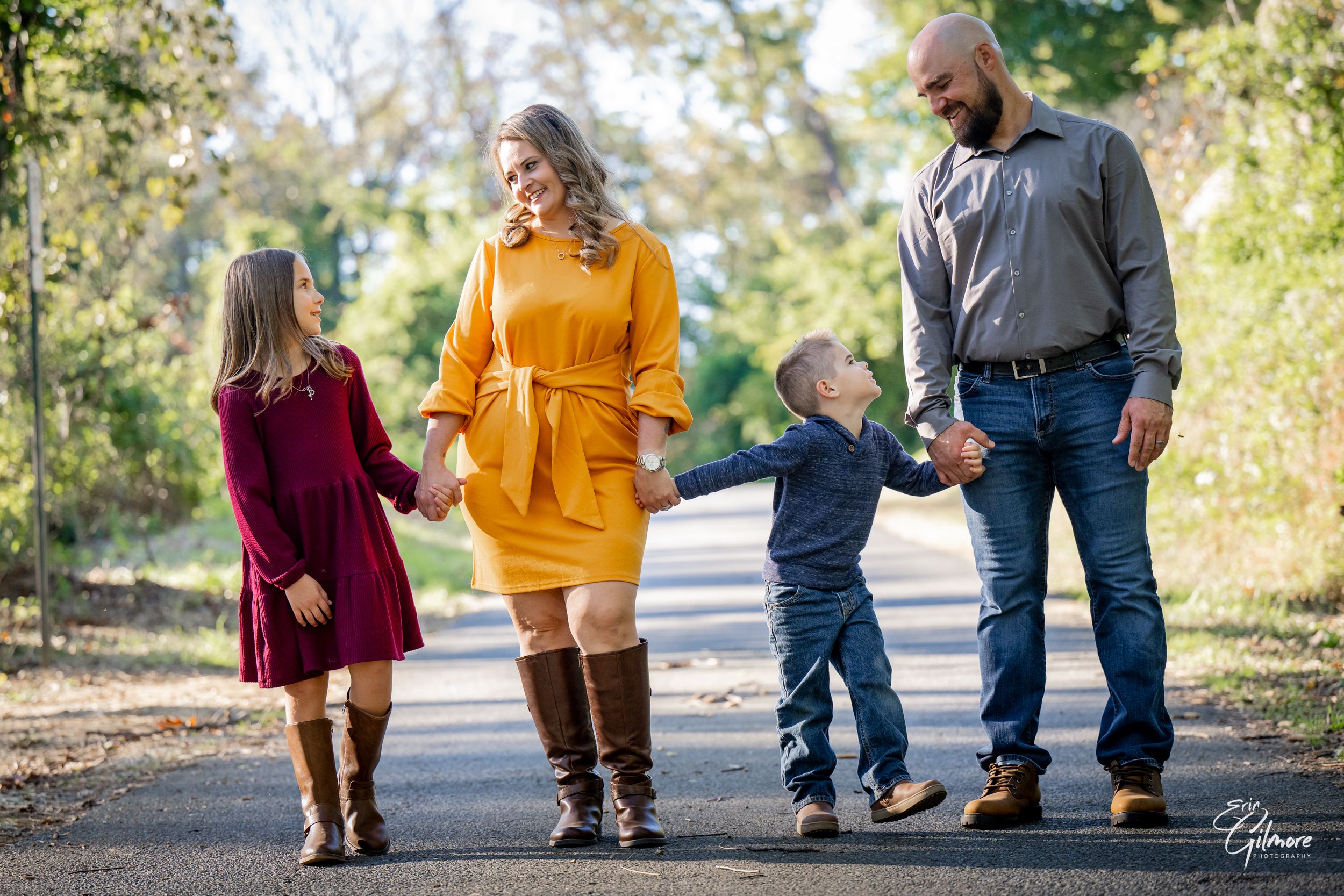 A happy family of four walks hand-in-hand on a tree-lined path in autumn, wearing warm fall colors.