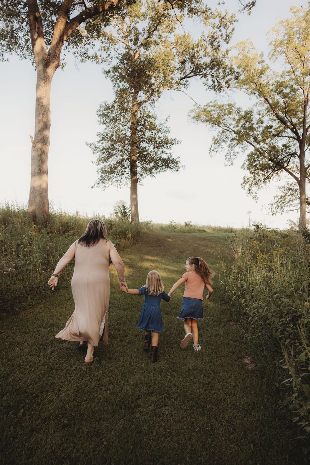 A mother and two daughters walk hand-in-hand through a lush green park path surrounded by tall trees at sunset.