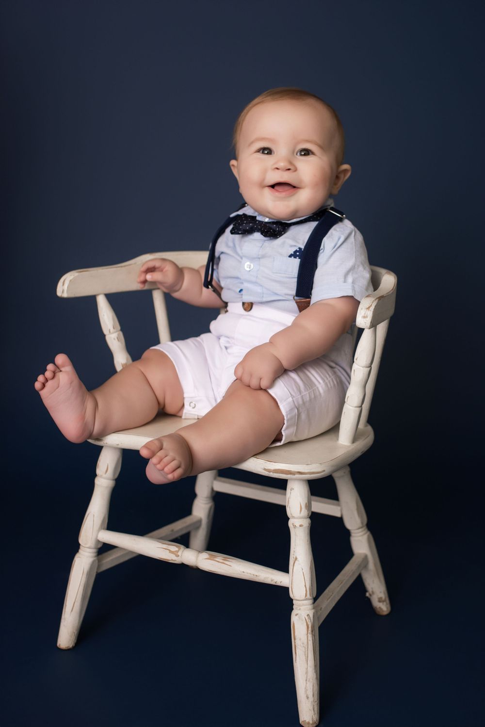Baby in white outfit sitting on vintage white wooden chair against navy backdrop.
