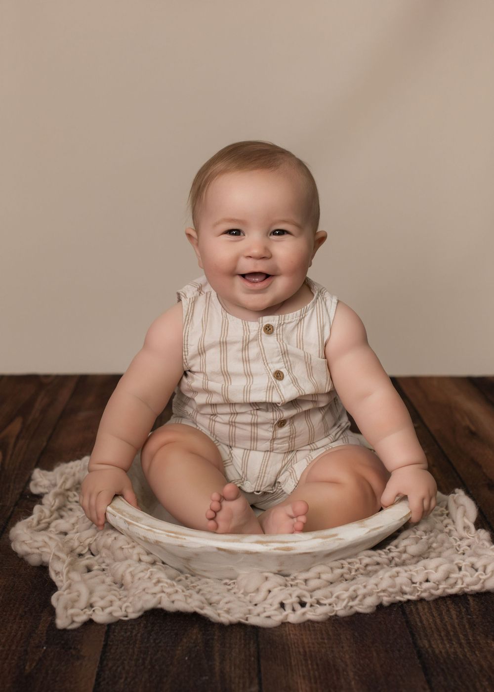 Baby in striped romper sitting on wooden surface with cream knit blanket.