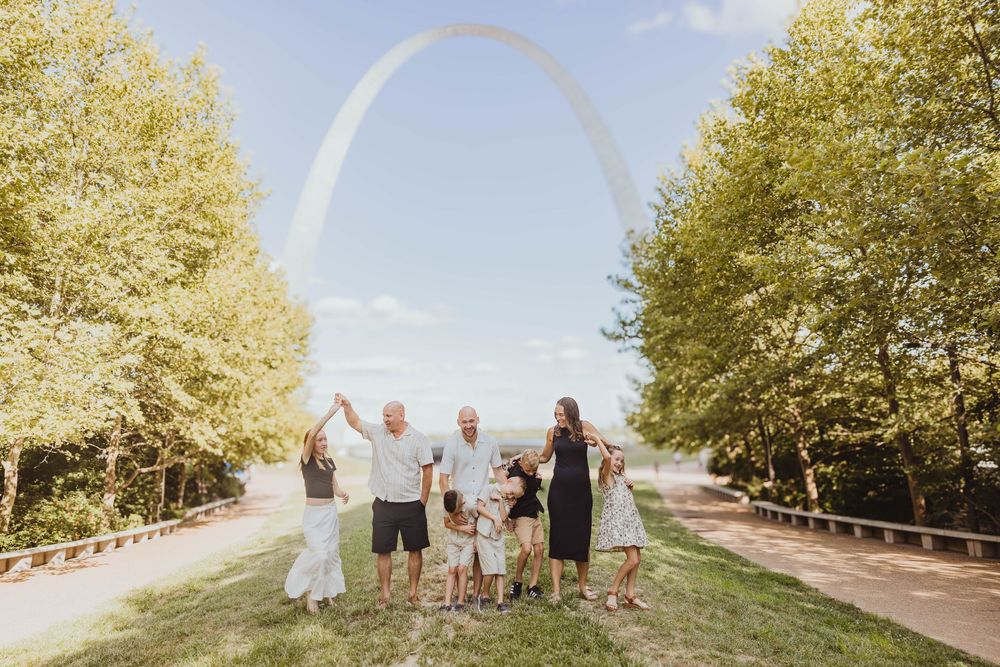 Family posing together in front of the Gateway Arch in St. Louis on a sunny day surrounded by green trees.